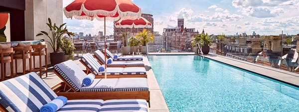 The rooftop pool and bar at The Berkeley with blue and white striped sun loungers, shaders and a view on Hyde Park and the London rooftops on the right, and the bar with bar stools on the left.